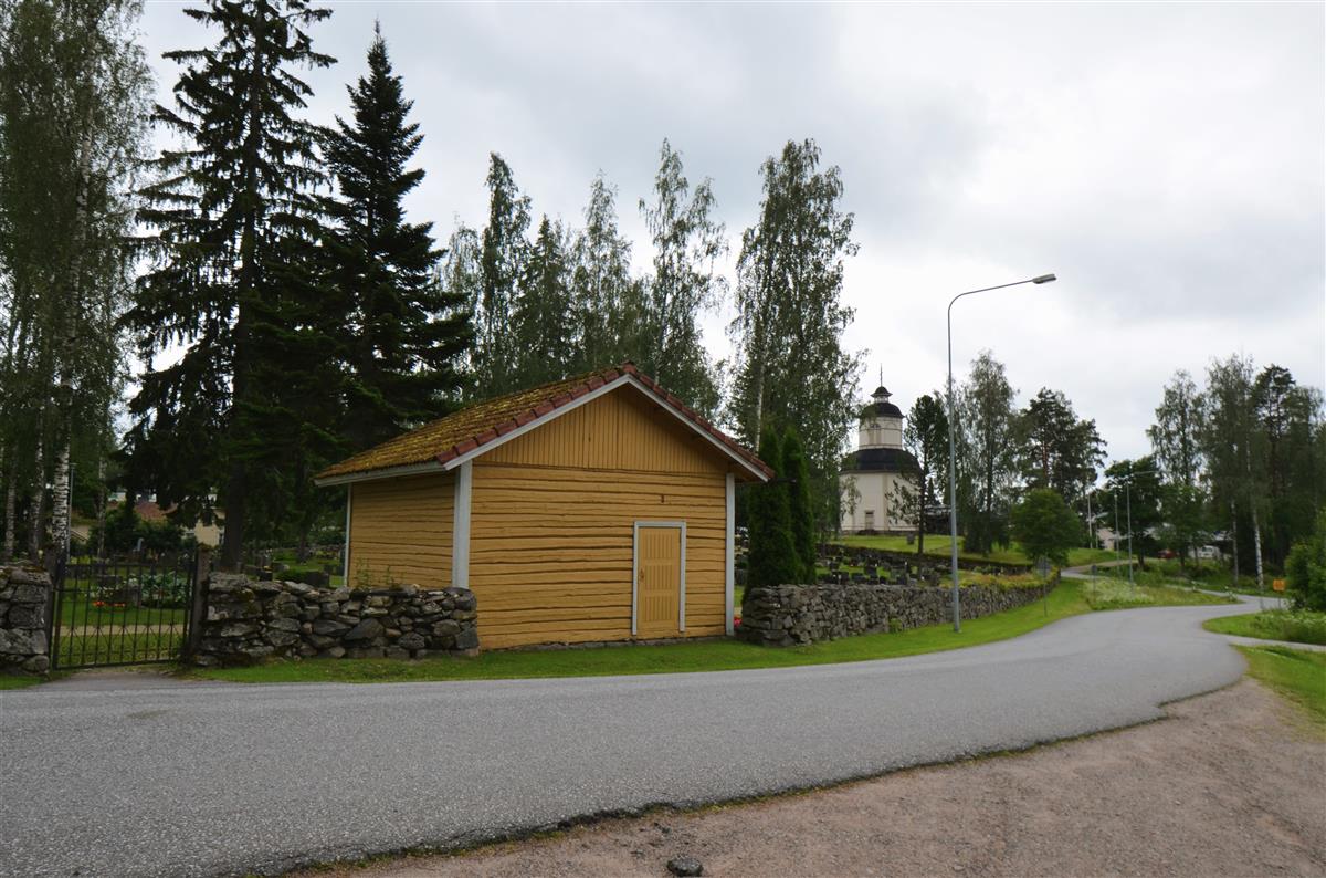 Hautausmaan leikkuuhuone Paimensaarentien yli, pohjoisesta päin kuvattuna. Taustalla Savitaipaleen kirkon kellotapuli.&nbsp;(Päivi Makkonen&nbsp;2025)